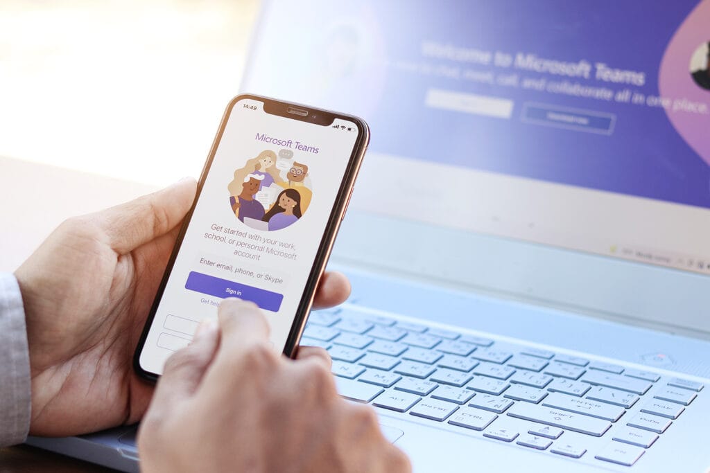 A person holding a phone in front of a laptop to show unified communications across multiple devices through Microsoft Teams.