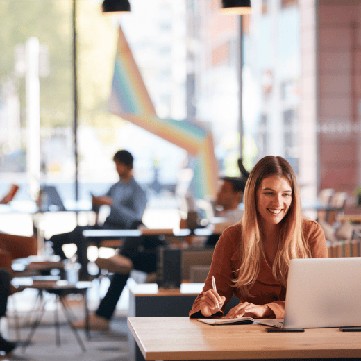 A woman in a coworking space on her laptop taking notes with pen and paper. She is smiling at her screen.