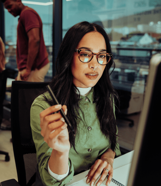 A woman in an office is looking at her screen. She holds a pen in her hand and is ready to take notes.