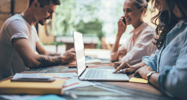 Three people at a co-working table. One is looking at his phone, one is looking at a laptop and the third one is taking a phone call.