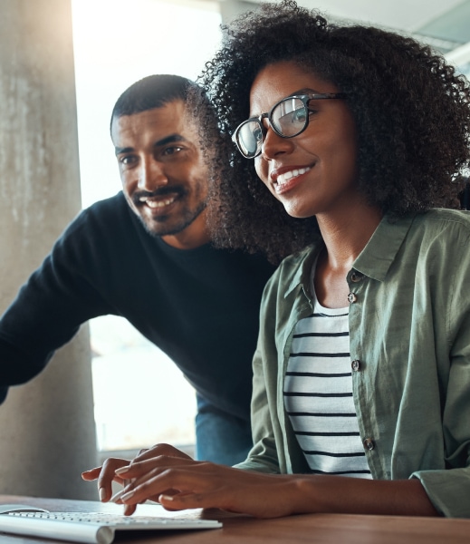 Two co-workers are looking at a computer screen. They’re both smiling.