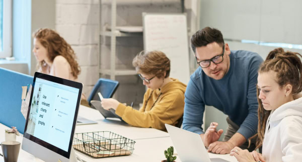 Coworkers in the office, two of them look at a laptop screen together, the other two look at their own screens.