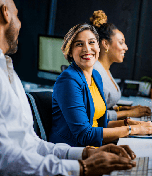 Three coworkers sitting in front of their computers. Two of them are smiling at each other.